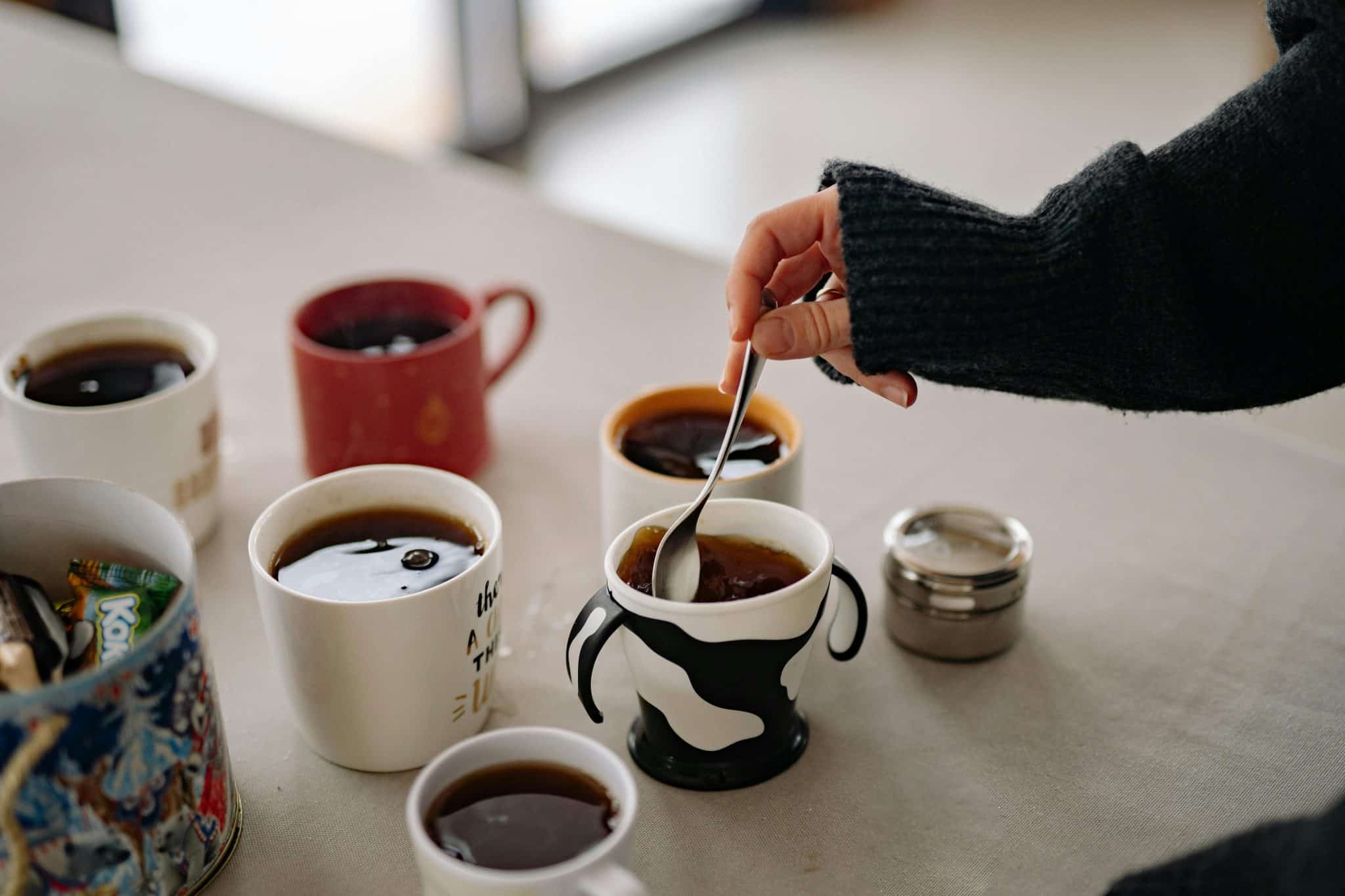 A top-down view of a hand stirring a cup of tea on a table filled with several other steaming mugs of tea in various colours and designs.