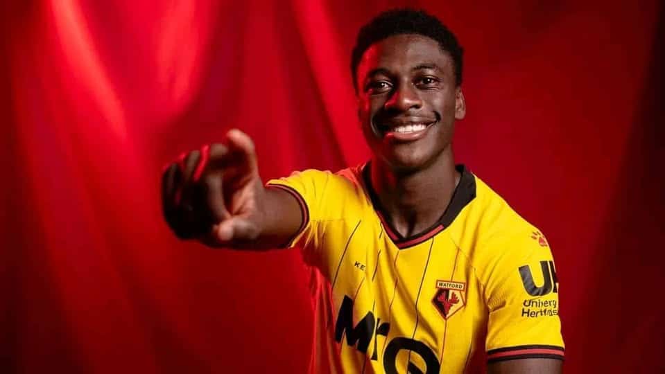 James Abankwah smiling and pointing toward the camera while wearing his yellow and black Watford football kit.