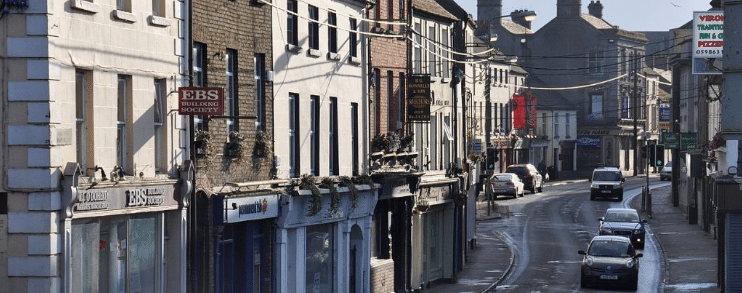 A sunlit street in Athy, Kildare, featuring traditional multi-story storefronts, local business signs like EBS, and a line of cars parked along the curb.