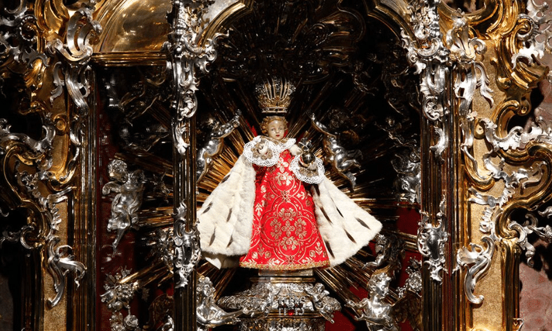 A small, ornate statue of the Infant Jesus of Prague wearing a red robe, a white fur-lined cape, and a golden crown, set against a highly decorative gold and silver altar.
