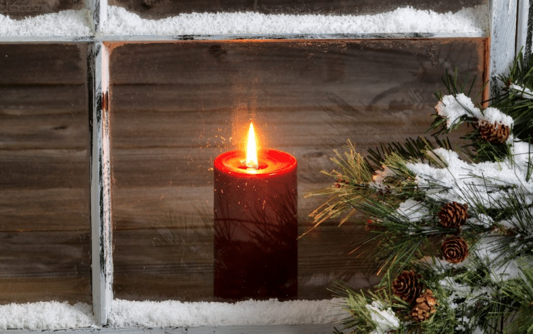 A thick red candle with a glowing flame sits behind a rustic, snow-dusted window pane next to a frosted evergreen branch.