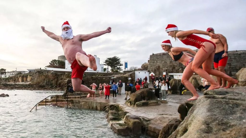 A man wearing a Santa hat and a fake white beard leaps enthusiastically into the cold sea from a rocky ledge during a Christmas Day swim.
