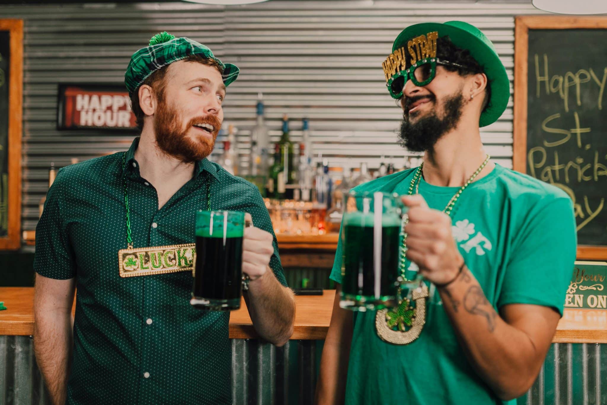 Two men in festive St. Patrick's Day gear stand at a bar holding mugs of green beer, smiling at each other.