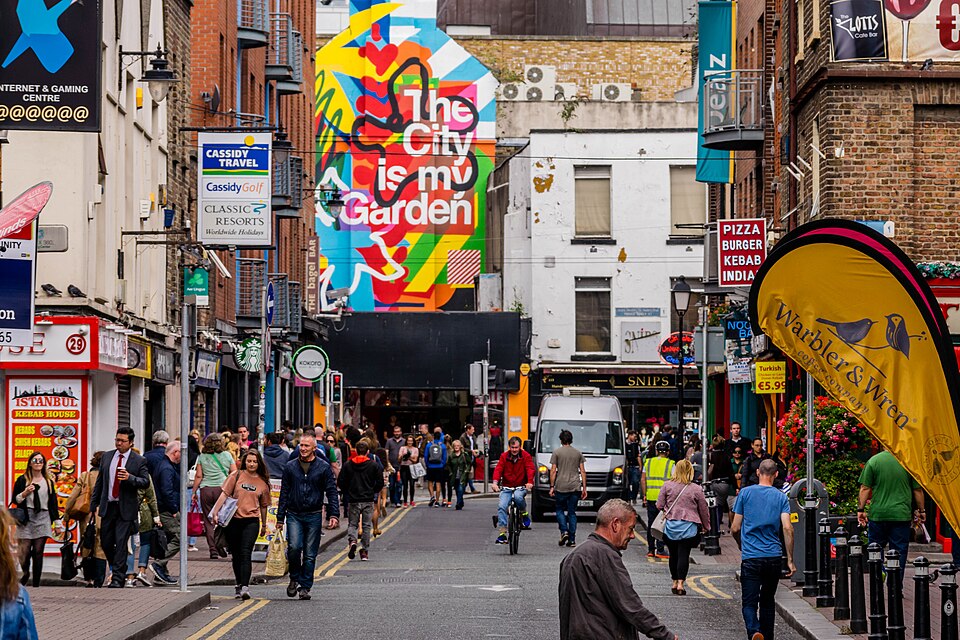 A busy, pedestrian-filled street in Dublin City featuring colourful shopfronts, various business signs, and a large, vibrant mural that reads "The City is my Garden."