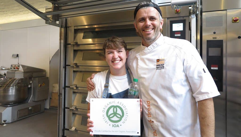 Eoin Cluskey (right), founder of Bread 41, smiling alongside a colleague who is holding a certification from the Irish Organic Association in their bakery.