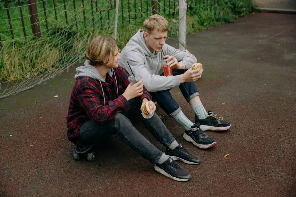 Two young people sitting on a skateboard eating burgers.
