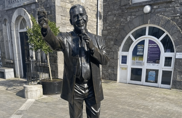 A bronze statue of Irish singer Joe Dolan holding a microphone, located in front of a stone building in Mullingar.
