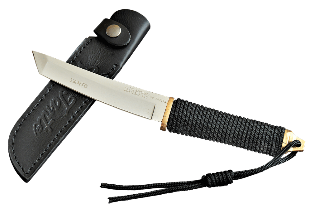 A sharp, modern Tanto-style knife with a black cord-wrapped handle lies next to its black leather sheath.