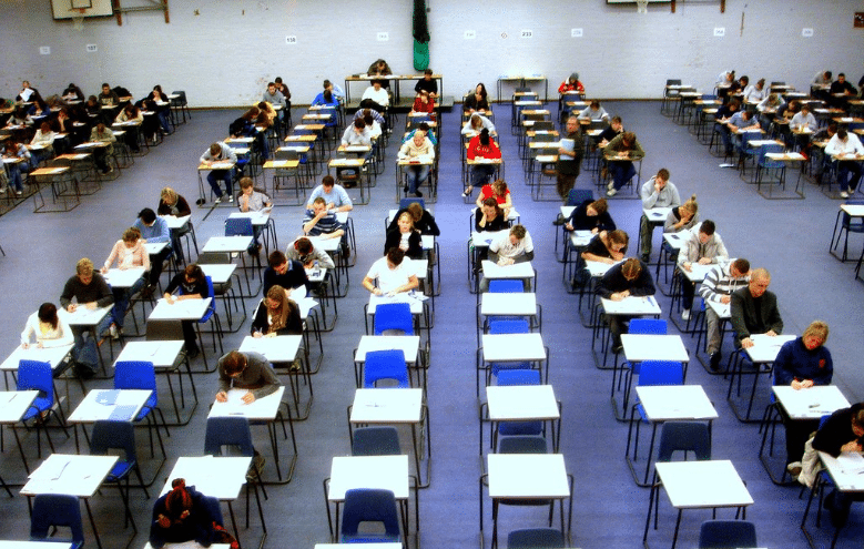 A high-angle view of students sitting at individual desks in a large, blue-floored hall while taking a formal examination.