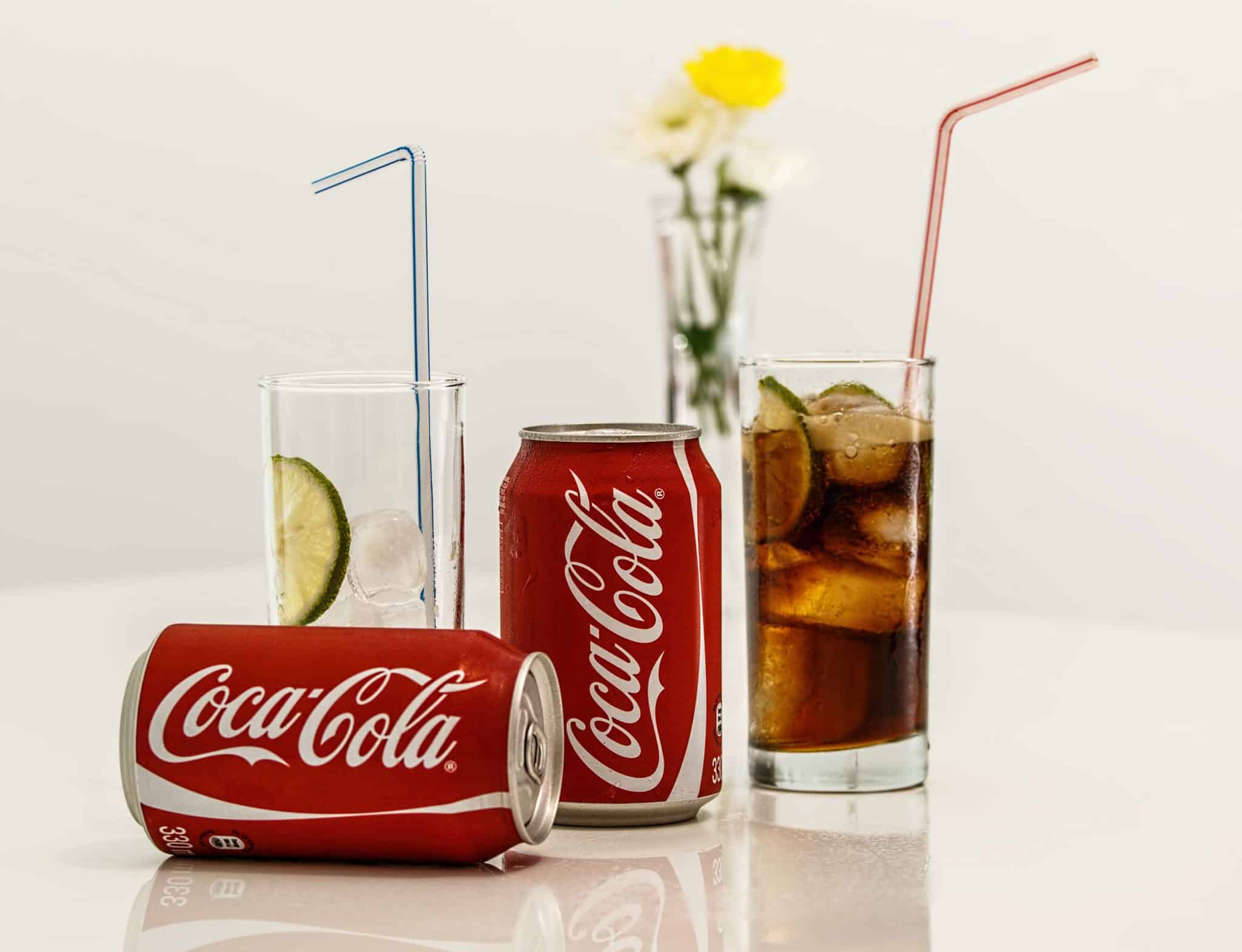 A Coca-Cola can lies on its side next to a standing can and a glass of cola with ice, lime, and a straw, all set against a white background with a small vase of flowers in the distance.