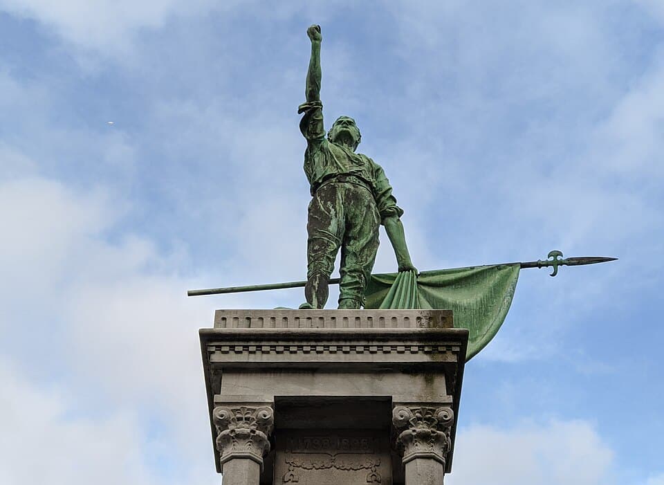 A low-angle shot of the 1798 Rebellion monument in New Ross, showing a bronze statue of a man holding a pike and flag against a cloudy sky.