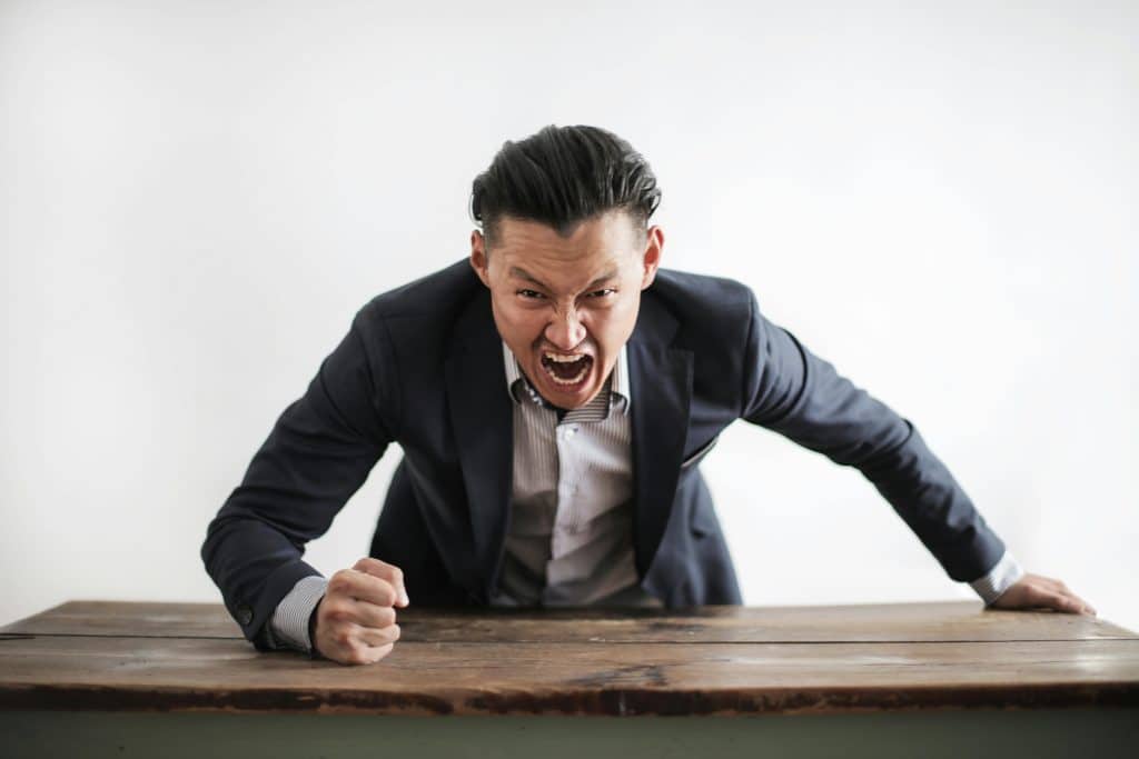 A man in a dark suit leaning over a wooden table with a clenched fist and an angry, shouting facial expression.