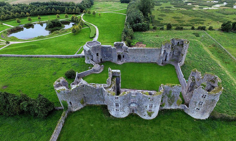 An aerial view of the stone ruins of Roscommon Castle, showing its interior green courtyard and surrounding parkland with a small lake.