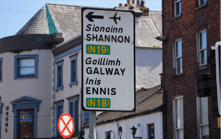 A close-up of a white directional road sign pointing toward Shannon, Galway, and Ennis, situated in front of urban buildings.