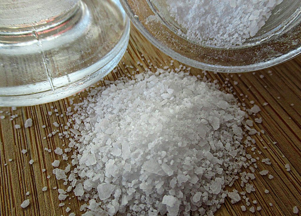 A pile of coarse white sea salt crystals scattered across a wooden surface, with a glass salt cellar visible in the background.