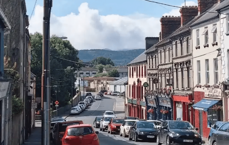 A view looking down a narrow, sloping street in Tipperary Town, lined with colourful shopfronts and a long row of parked cars leading toward distant green hills.