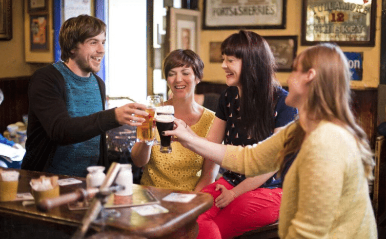A group of friends laughing and clinking glasses of beer and stout together over a wooden table in a traditional Irish pub.