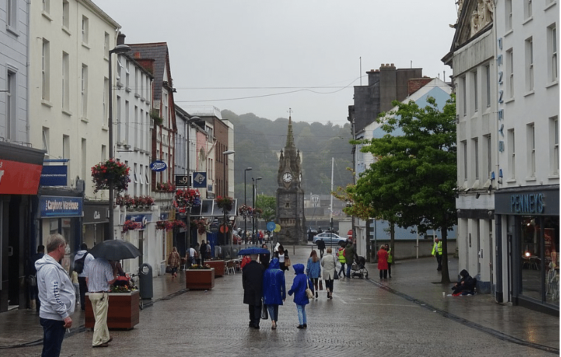 Pedestrians with umbrellas walk down a damp, paved shopping street in Waterford toward a distant stone clock tower under a cloudy sky.