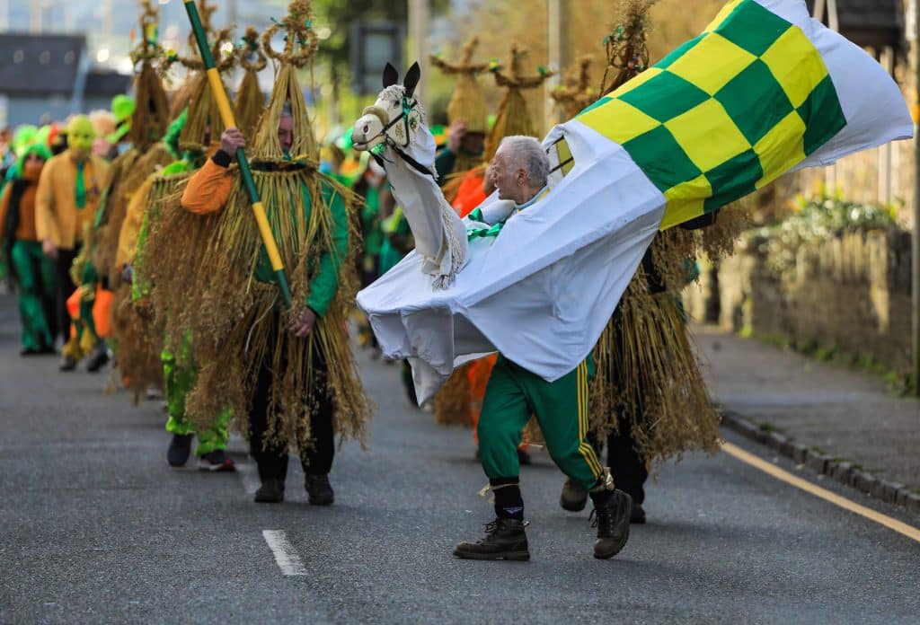 A parade of Wren Boys wearing tall, conical straw masks and suits, with one participant in the foreground wearing a hobby horse costume featuring a checkered green and yellow flag.