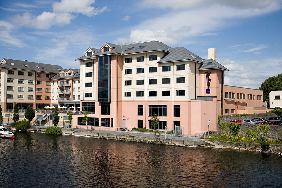 A multi-storey, peach and cream-coloured hotel building situated directly along the banks of the River Shannon in Athlone, Ireland, under a bright, partly cloudy sky.