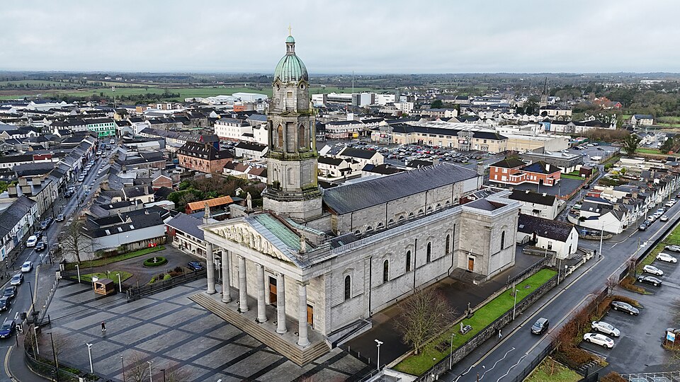 An aerial view of St Mel's Cathedral in Longford, Ireland, featuring its prominent neoclassical stone facade with large columns and a tall, green-domed bell tower, surrounded by the town's buildings and streets under an overcast sky.