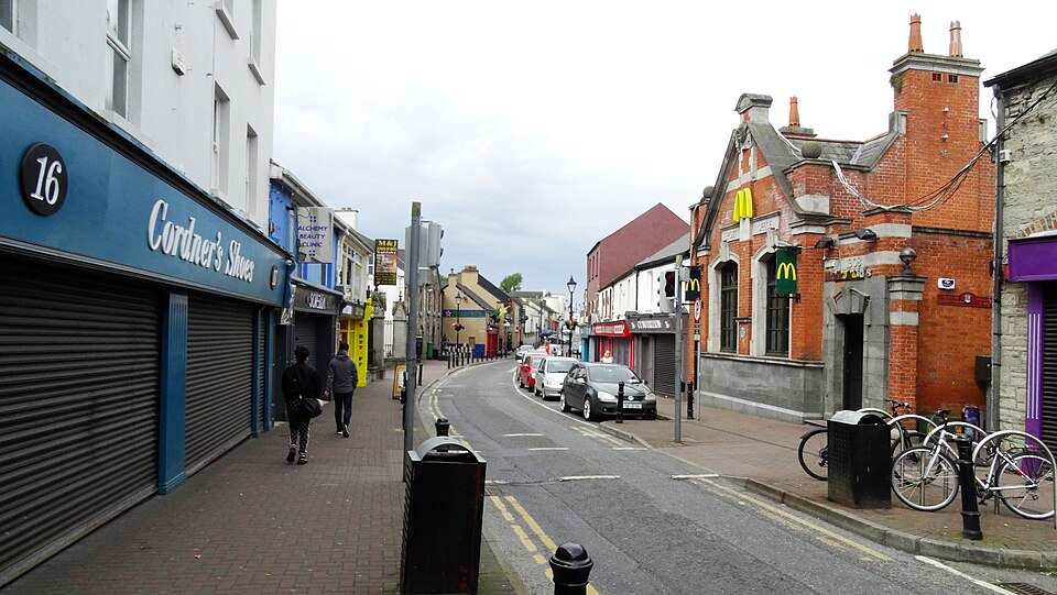 A view looking down Trimgate Street in Navan, Ireland, featuring a mix of traditional brick buildings and modern storefronts with closed shutters, with several parked cars along the narrow road and bicycles secured to a rack in the foreground.