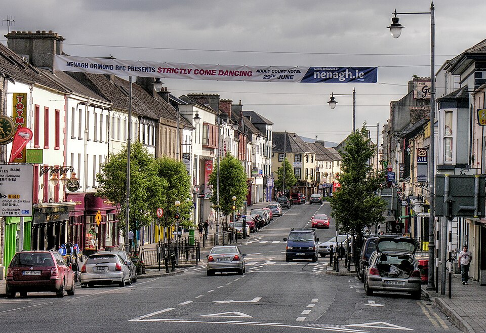 A busy street scene in Nenagh town centre, Ireland, featuring rows of traditional shops and businesses with a large event banner stretched across the road, while cars travel through and park along the kerbside under an overcast sky.