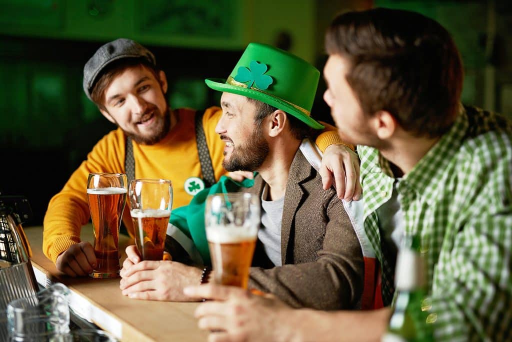 Three men at a bar celebrating St. Patrick's Day, wearing green hats and shamrock accessories while enjoying glasses of beer.