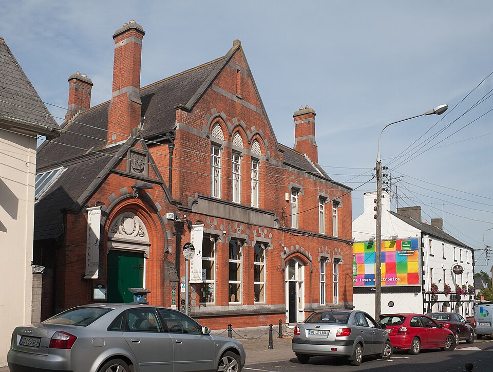 A historic red-brick building with tall chimneys and arched windows on a street in Portlaoise, featuring parked cars in the foreground and a modern, colourful billboard in the background.