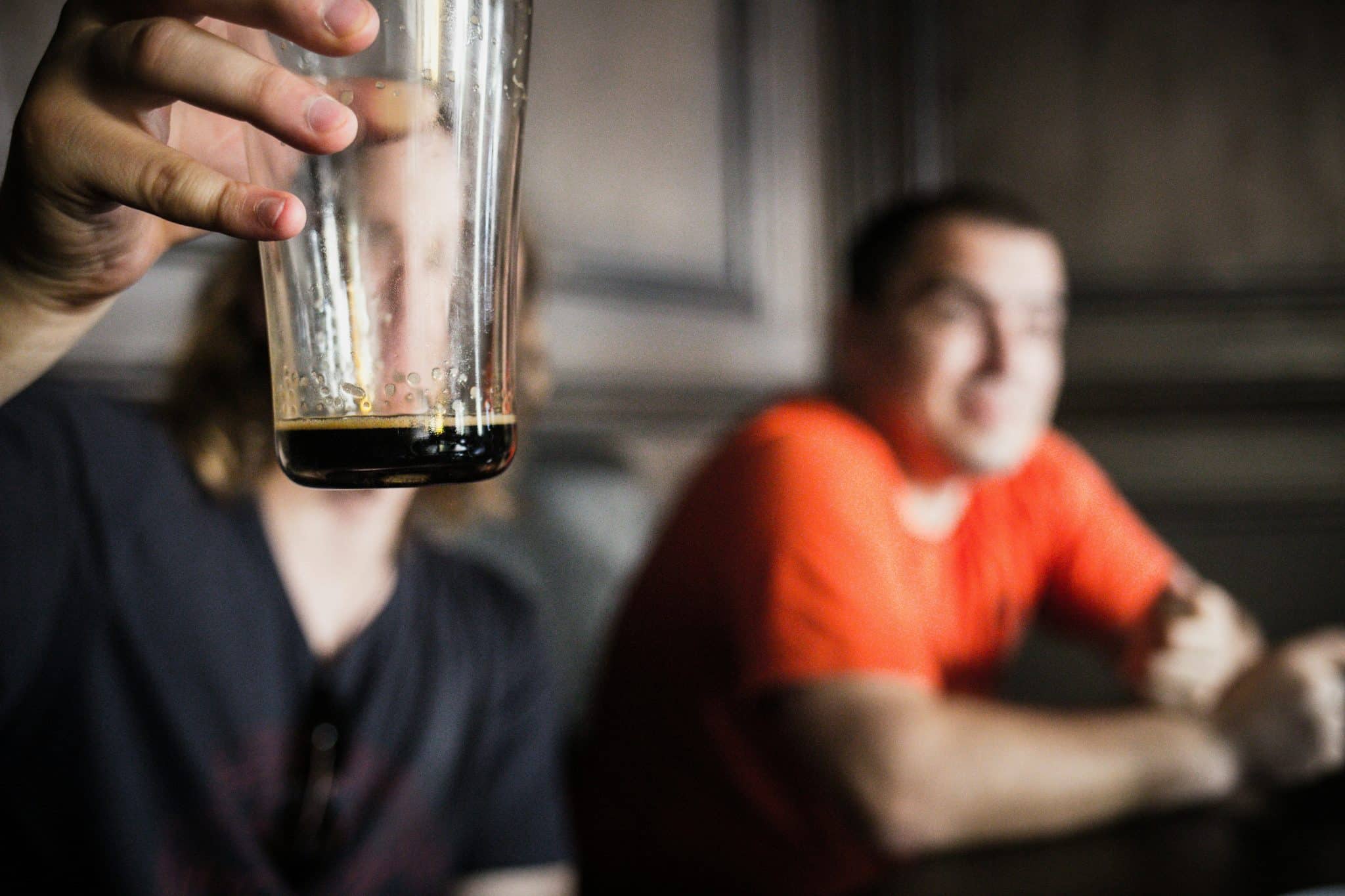 Man holding up a near empty glass with a man blurred in the background.