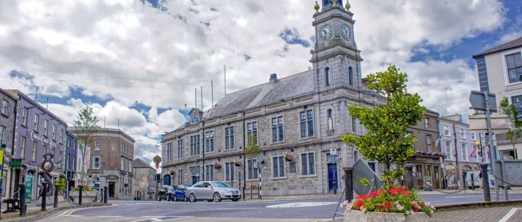 A panoramic view of Tuam town square featuring the historic stone Town Hall with its prominent clock tower, set against a bright, cloudy sky with local shops and a flower planter in the foreground.