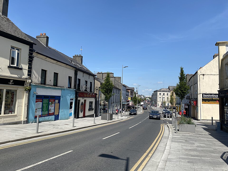 A view of High Street in Tullamore, Ireland, showing a wide, paved road with modern street lighting and young trees, flanked by traditional two-storey commercial buildings under a clear blue sky.